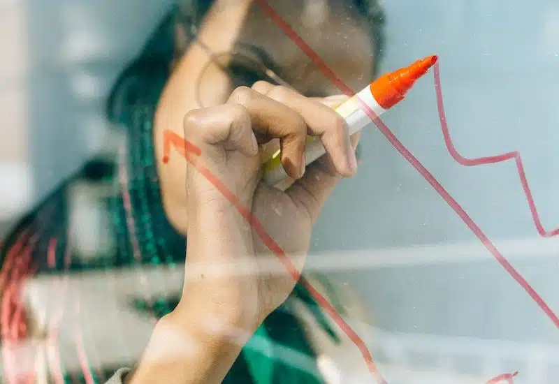 Woman in a Beige Coat Writing on a Glass Panel Using a Whiteboard Marker